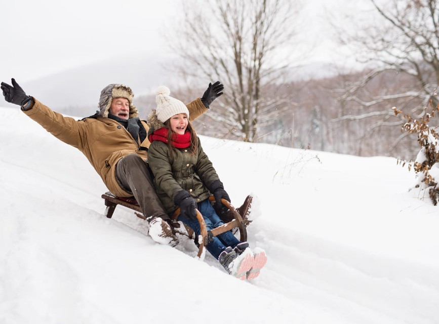 Vater und Kind rodeln fröhlich einen schneebedeckten Hang hinunter und genießen den Winterspaß im Familienurlaub © Shutterstock