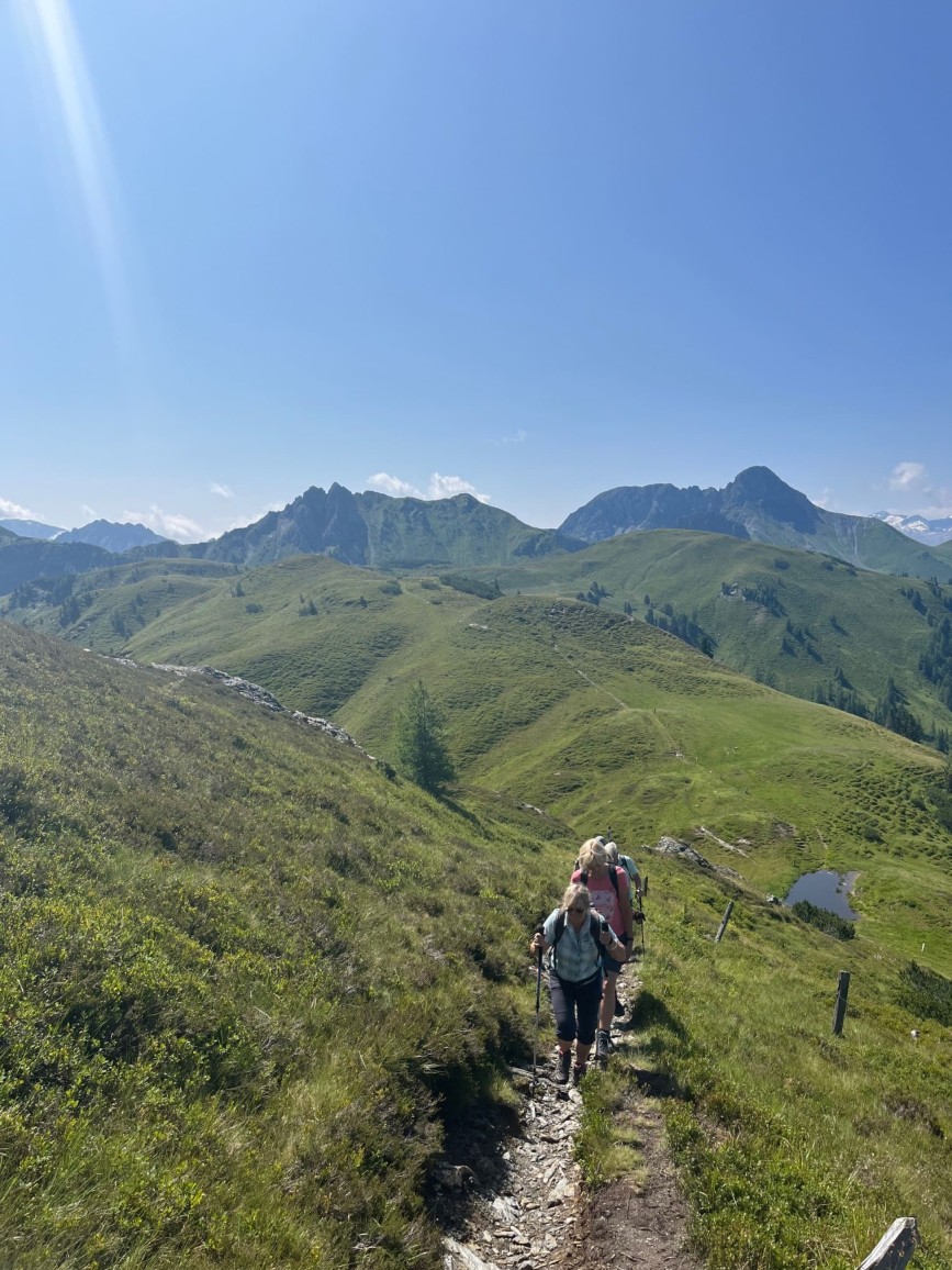 Hikers on a mountain trail with rolling alpine meadows, mountain peaks and a small alpine pond in the background